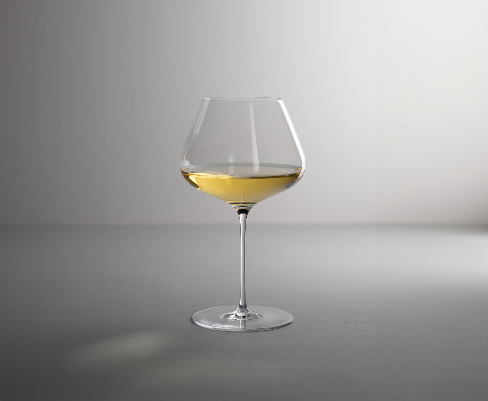 Das mit Weißwein gefüllte Denk'Art Balance Weinglas steht vor einem grauen Hintergrund. Der Lichteinfall lässt den Wein leuchten.