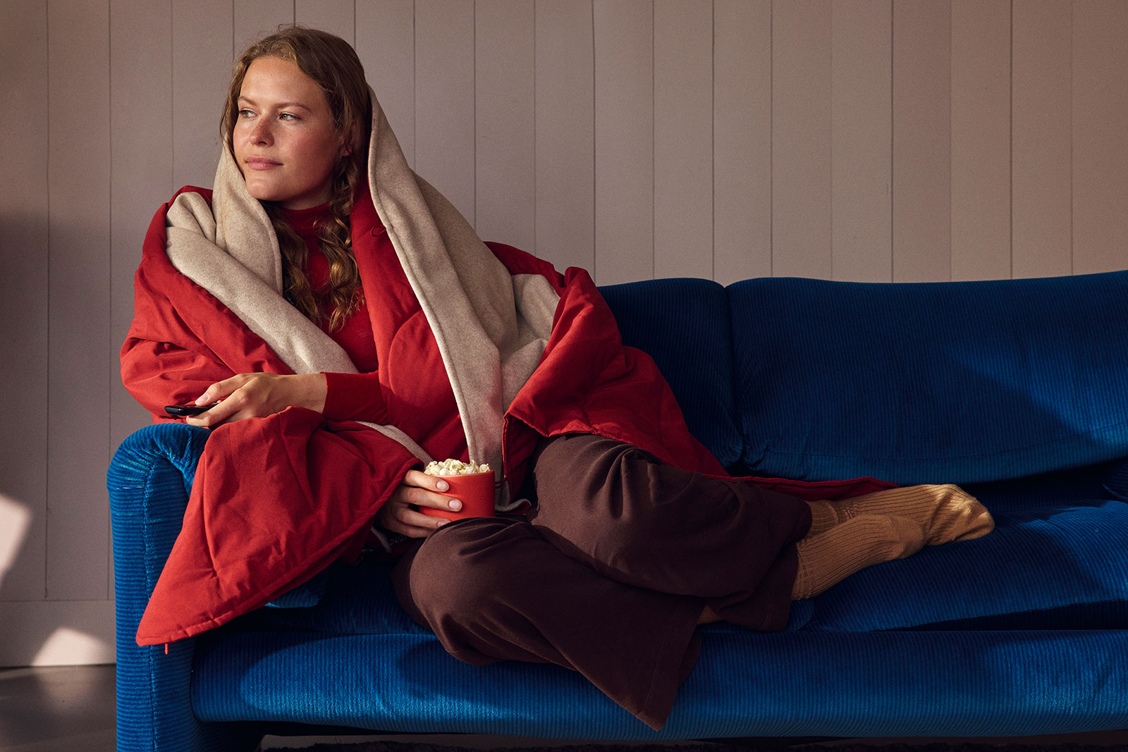 Eine Frau hat die Huge Hug Heizdecke Eart Red um sich gewickelt und sitzt auf einem blauen Sofa.