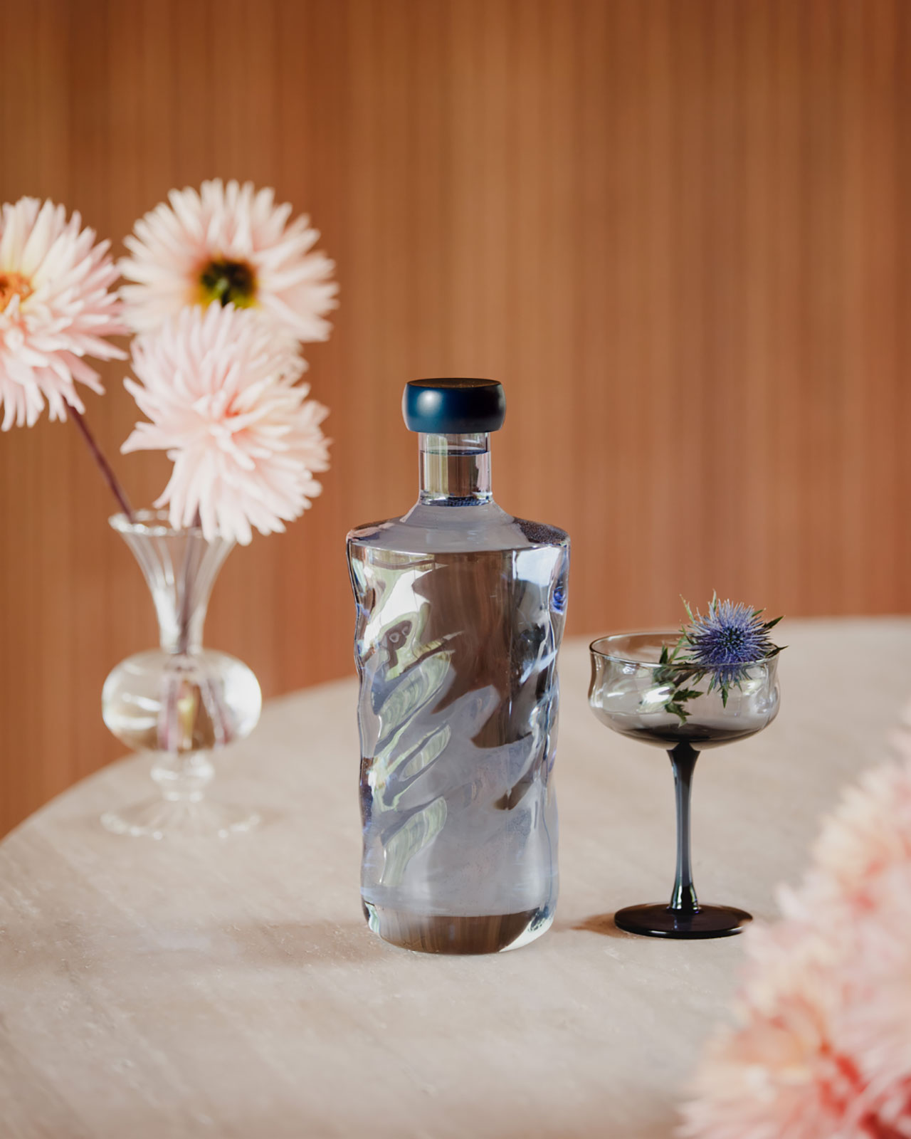 Auf einem gedeckten Tisch steht eine dunkelblaue Glasflasche neben einem Coupe-Glas der gleichen Farbe. In dem Glas befindet sich eine blaue Distel.