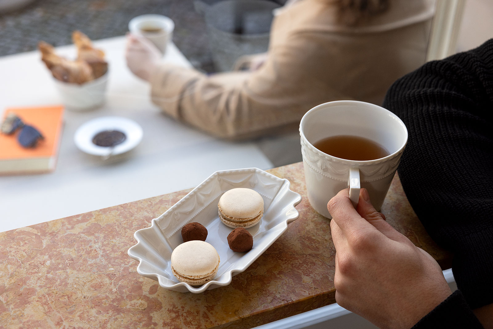 In einem Café hält jemand die Kurland weiß Mug in der Hand. Im Hintergrund steht die Kurland weiß Currywurstschale, gefüllt mit Macrons und Pralinen.