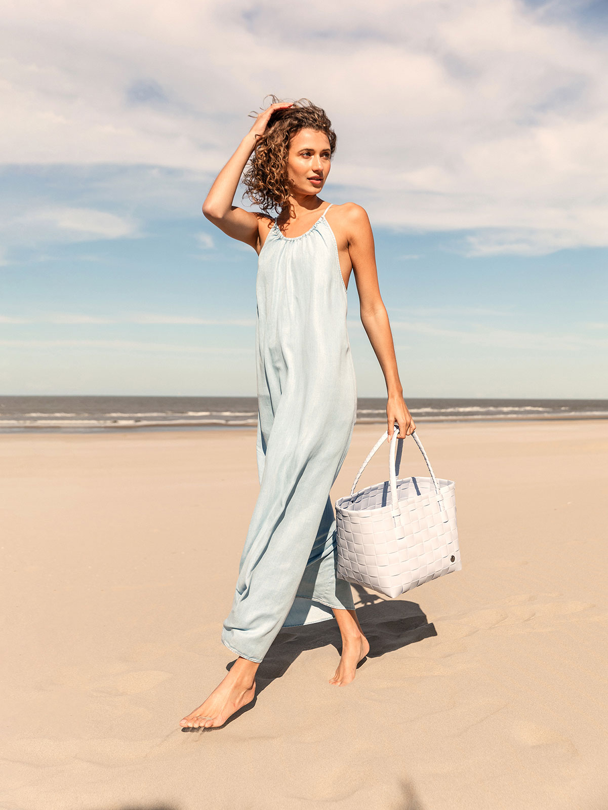 Eine Frau im langen hellblauen Kleid trägt den Paris Shopper icy mist den Strand entlang.
