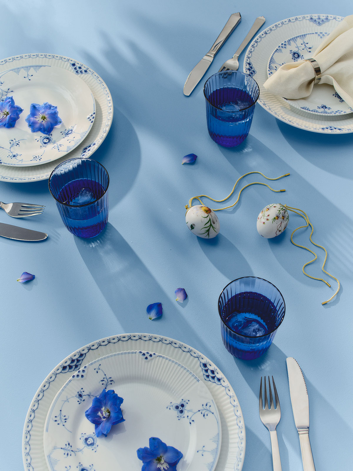 Auf einer blauen Tischdecke, zwischen blau dekoriertem Porzellangeschirr, liegt das Osterei Große Kamille 2026 von Royal Copenhagen.