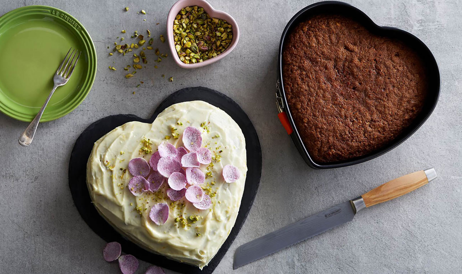 Auf einer mamornen Arbeitsfläche stehen die Herz-Springform von Le Creuset und ein Kuchen, der mit ihr gebacken wurde. Dieser ist romantisch verziert mit kandierten Rosenblättern.