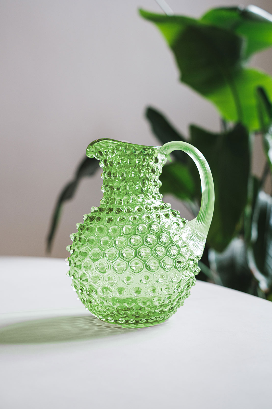 Auf einem weißen Tisch steht der Paris Hobnail Krug Light green von Anna von Lipa und zeigt, wie lebensfroh seine Farbe ist. Im Hintergrund befindet sich eine Pflanze mit großen, dunkelgrünen Blättern.