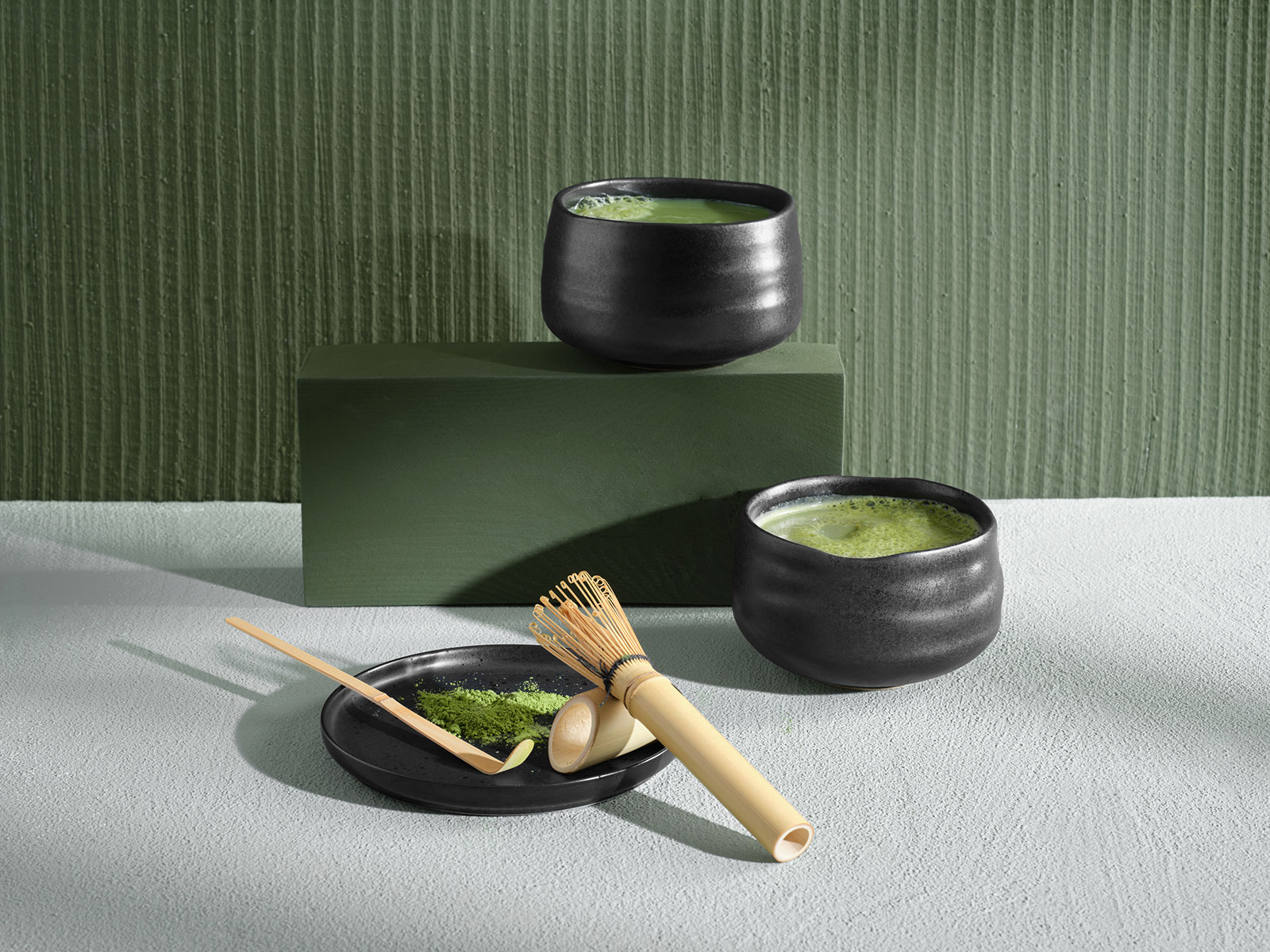 Vor einem grünen Hintergrund stehen zwei Moribana Matcha Schalen mit frisch zubereitetem Matcha. Im Vordergrund liegen auf einem Unterteller der Matcha Besen und der Löffel, die für die Zubereitung verwendet wurden.