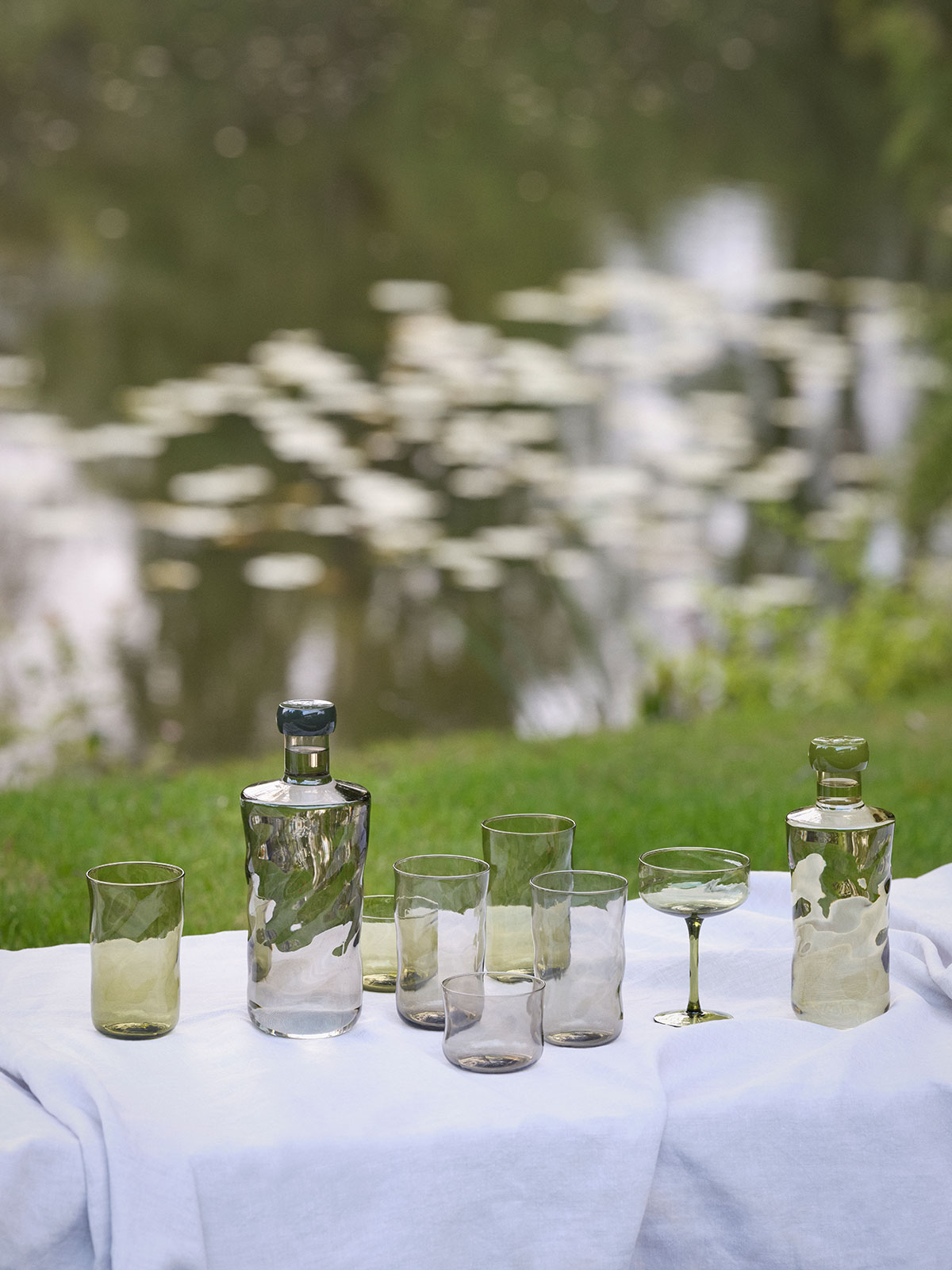 Auf einer weißen Tischdecke, ausgebreitet auf grünem Gras, stehen vermischt Wasserflaschen und Trinkgläser in grün und rauchgrau.