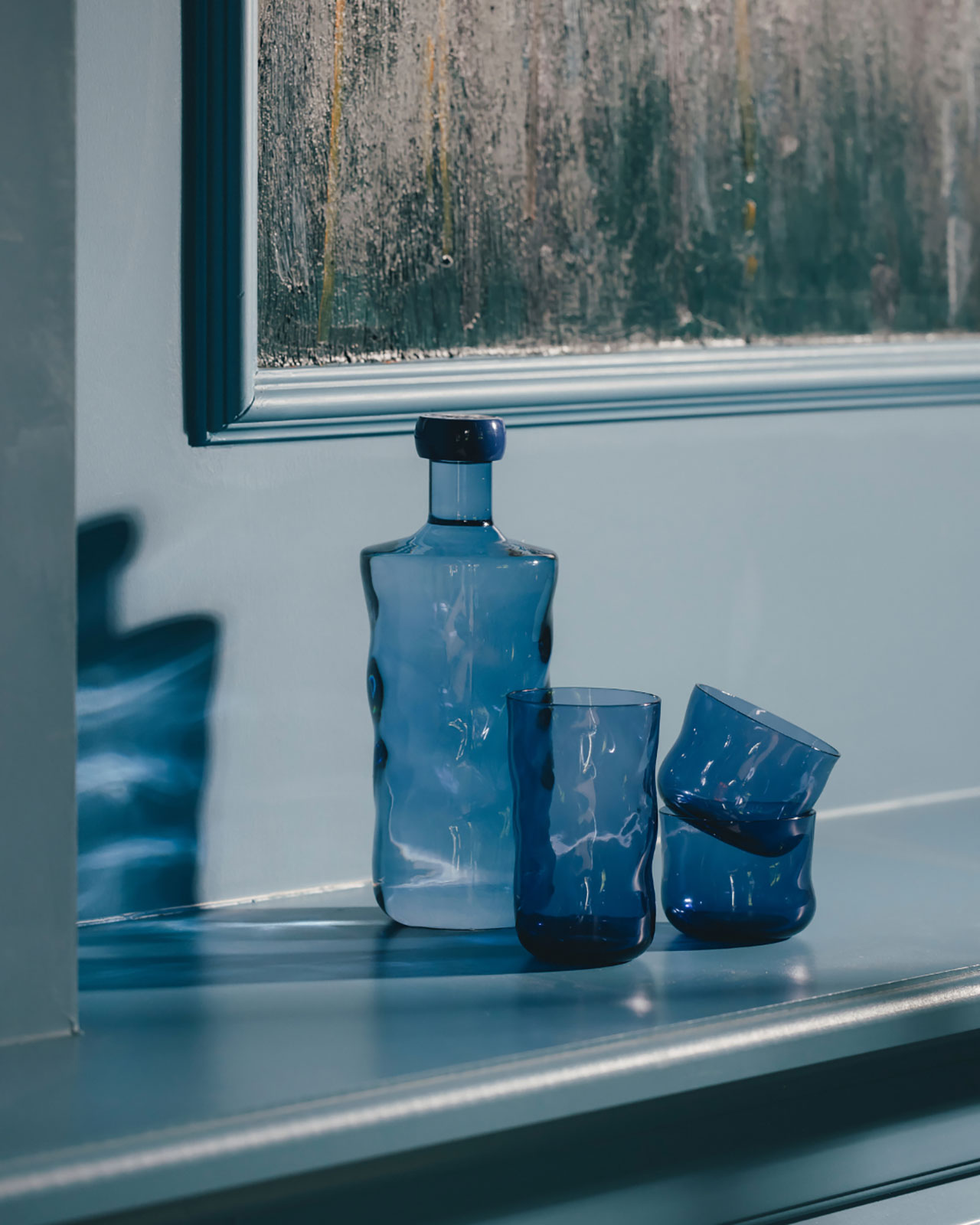 Vor einer blauen Wand steht auf einem Sideboard eine dunkelblaue Wasserflache neben mehreren ineinandergestapelten Trinkgläsern der gleichen Farbe.