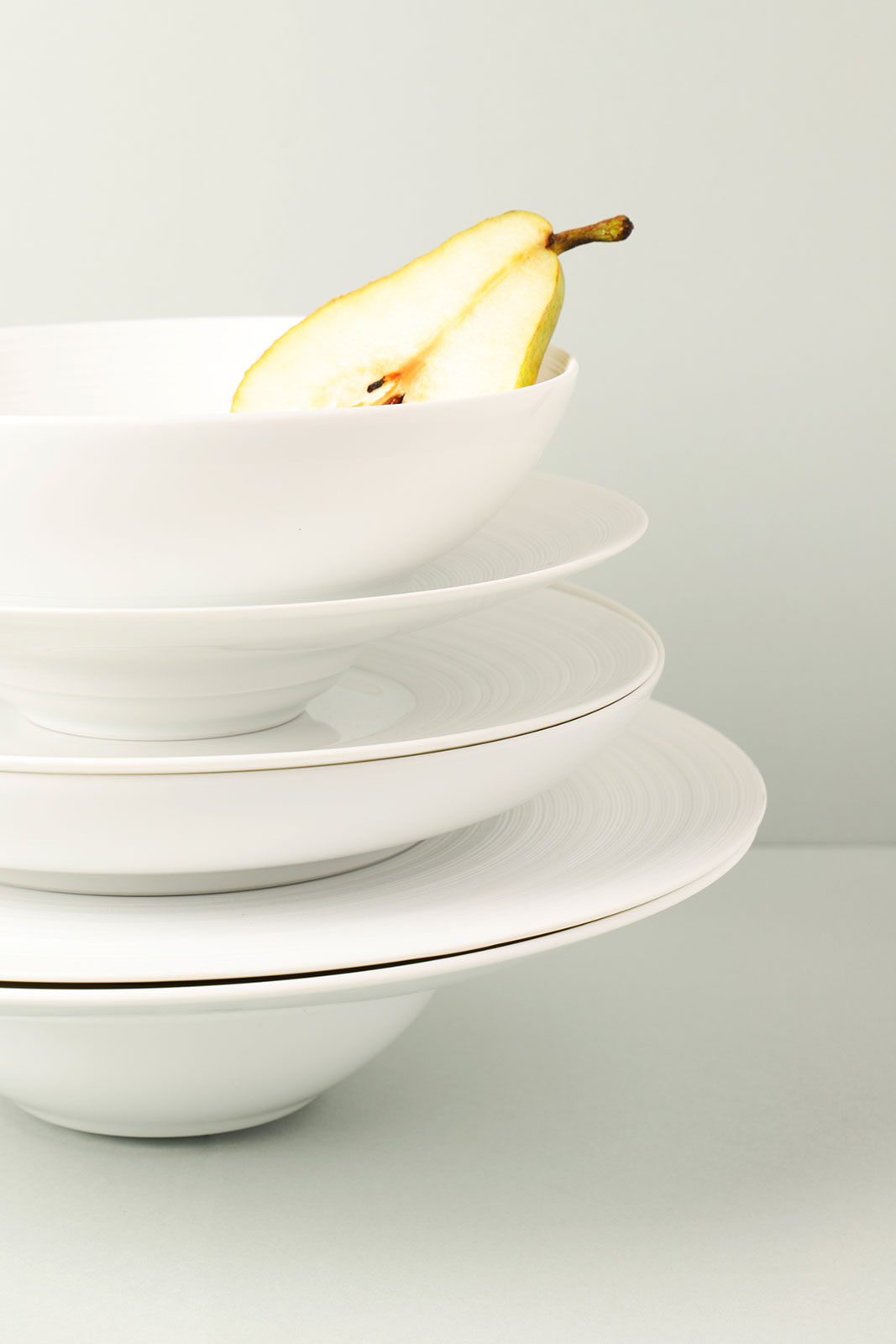 Verschiedene Teller und Schalen der Coquet Serie Hémisphère Satin white sind übereinander aufgestapelt. In der obersten Schale befindet sich eine aufgeschnittene Birne.