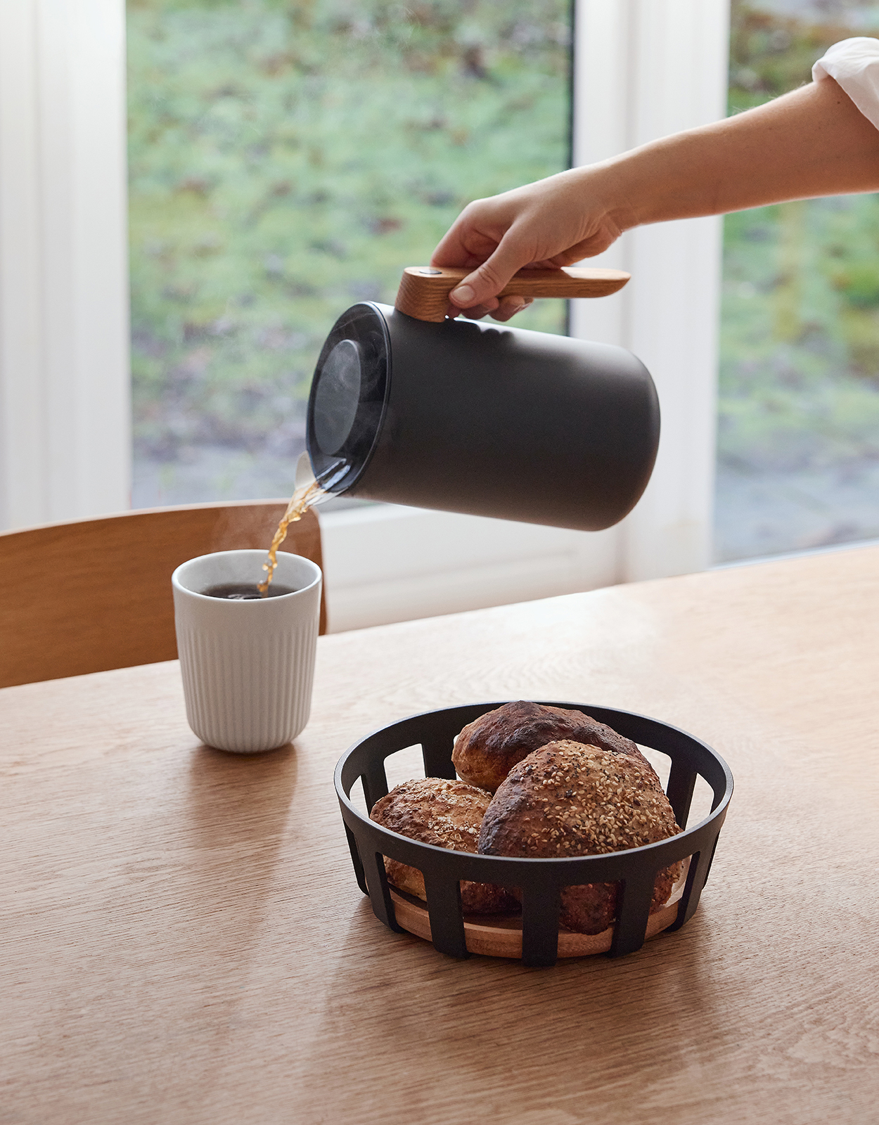 Ein Esstisch mit der Nordic Kitchen Isolierkanne, einem Brotkorm und einem Kaffeebecher, in den gerade Kaffee gegossen wird.