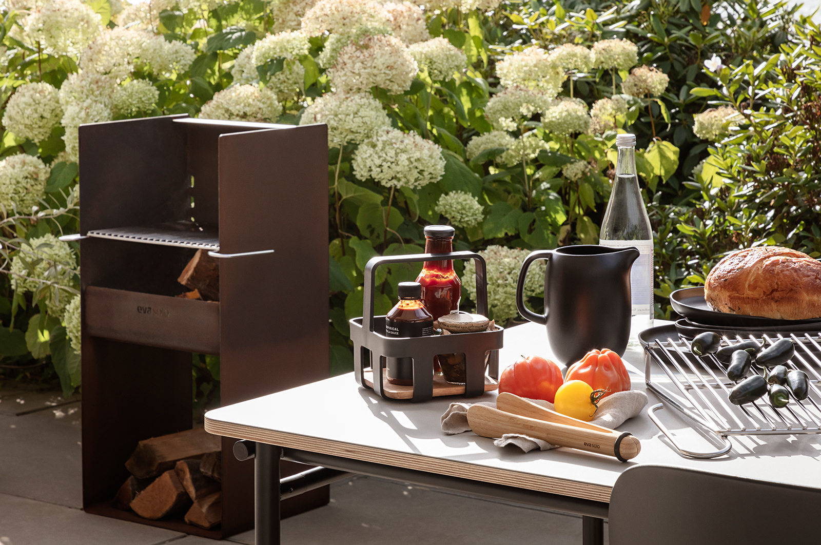 Ein Outdoortisch mit Grillutensilien. Im Zentrum des Bildes steht der Eva Trio Nordic Kitchen Caddy. Im Caddy stehen drei verschiedene BBQ Saucen.