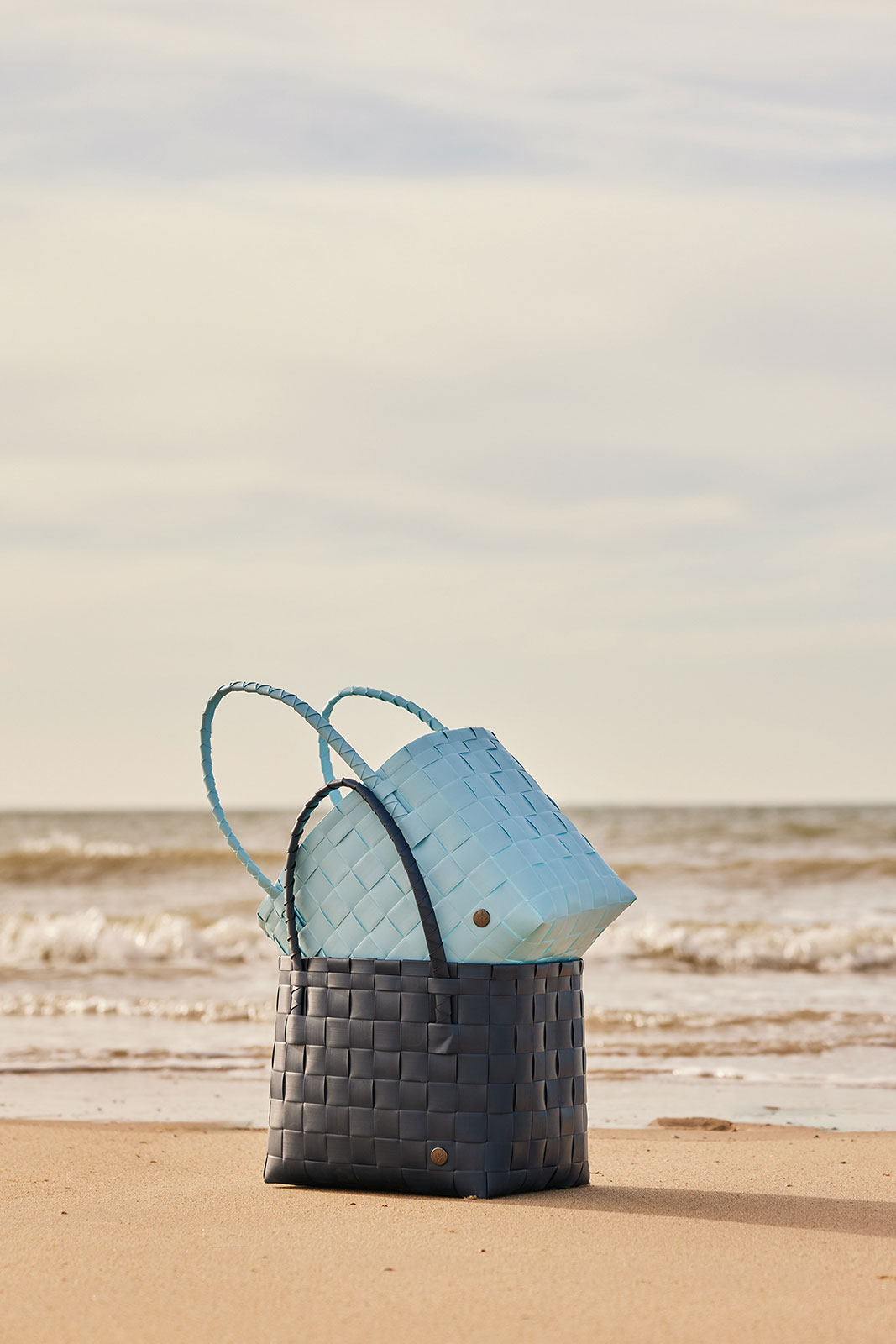 Der dunkelblaue Paris Shopper steht gemeinsam mit einem hellblauen Korb am Strand in der Brandung. Diese Körbe sind perfekt, um alles Nötige für einen Strandtag mitzunehmen!