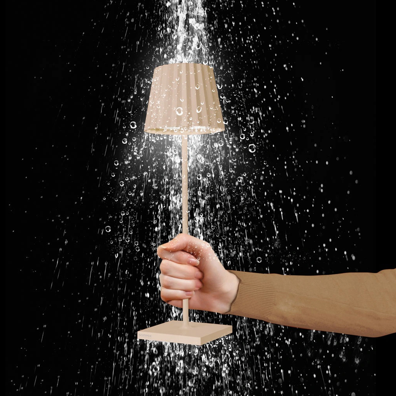 Weibliche Hand, die eine beige Akku-Tischleuchte in den Regen hält.