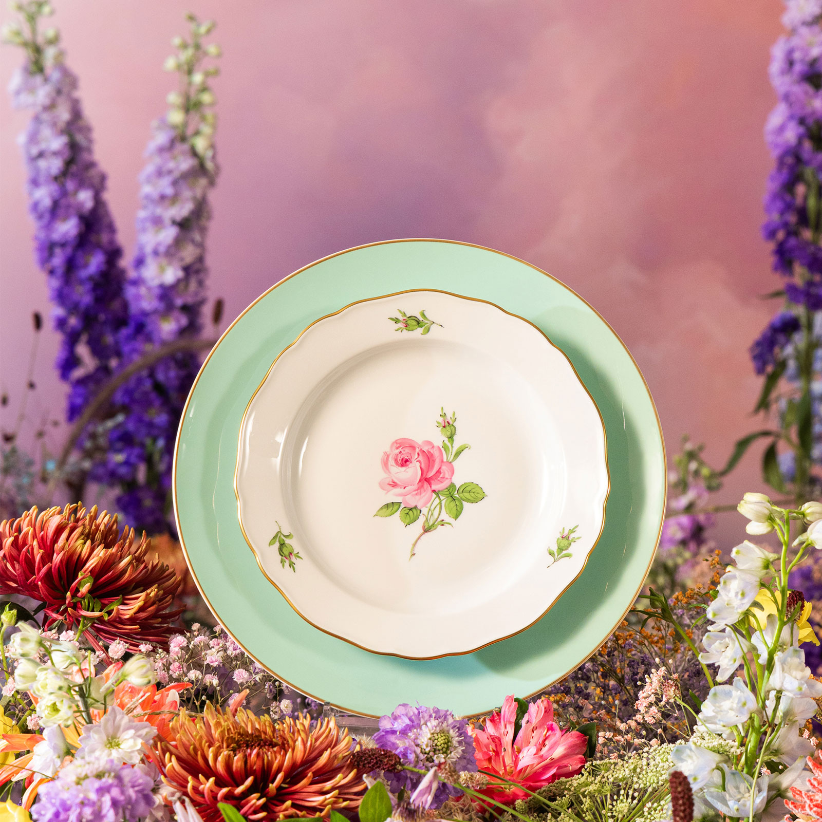 Vor einem Hintergrund aus arrangierten bunten Blumen steht ein Teller des Dekors Meissener Rose auf einem seladongrünen Urban Colors Speiseteller von Meissen.