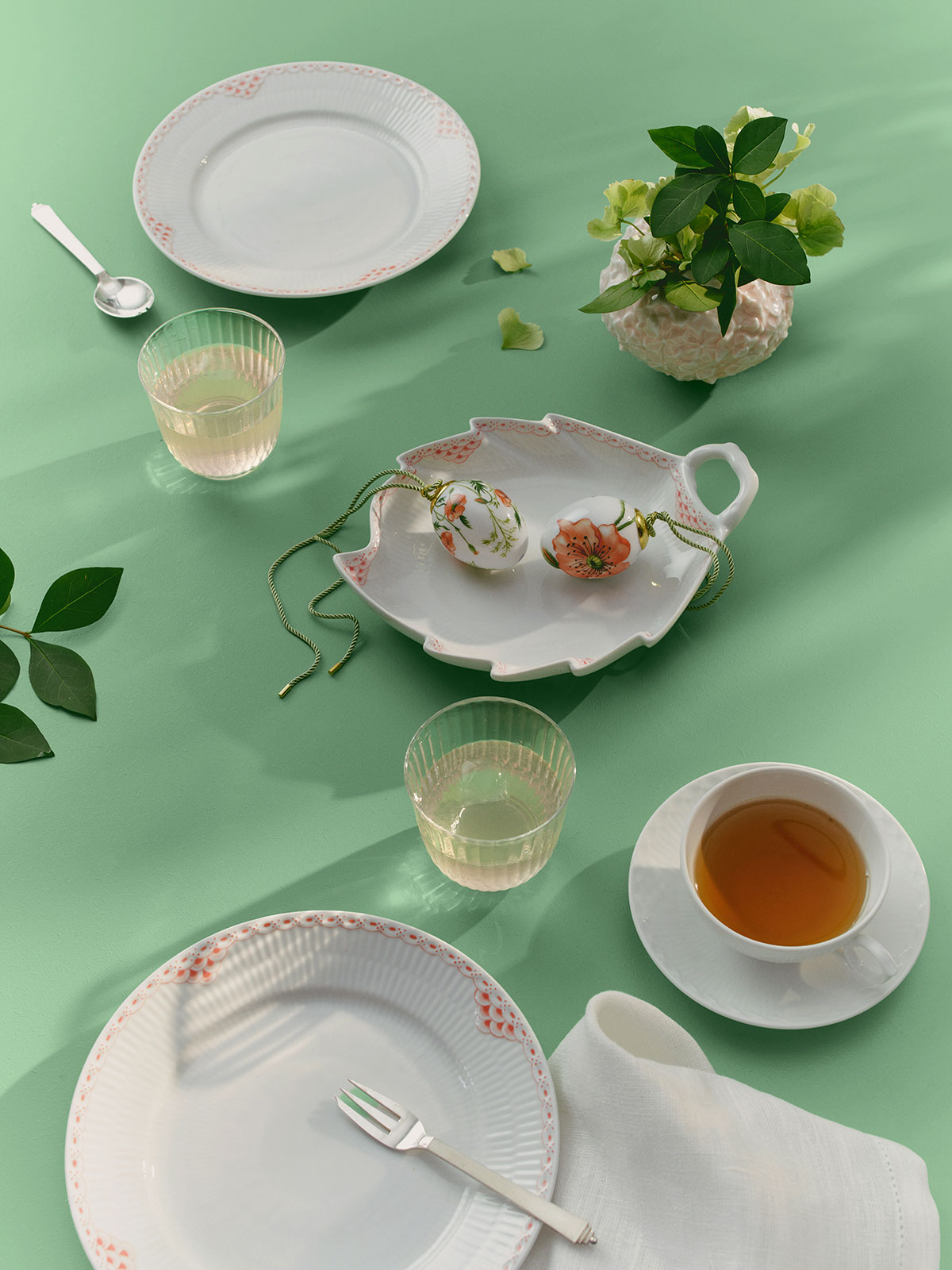 Auf einer pastellgrünen Tischdecke, zwischen weißem Porzellangeschirr und Gläsern mit Limonade, liegt das Osterei Große Kamille 2026 von Royal Copenhagen.