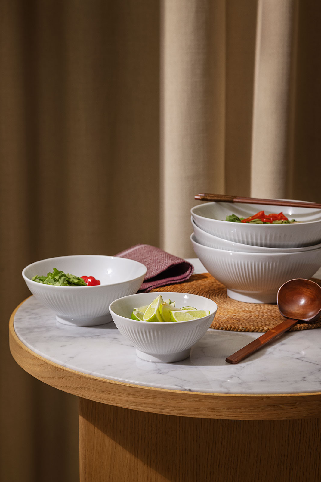Auf einem runden Tisch ist alles angerichtet, um Ramen-Nudeln zu genießen. In kleineren Legio Nova Schalen befinden sich Gemüse und Zitronenschlitze, um das Gericht zu verfeinern.