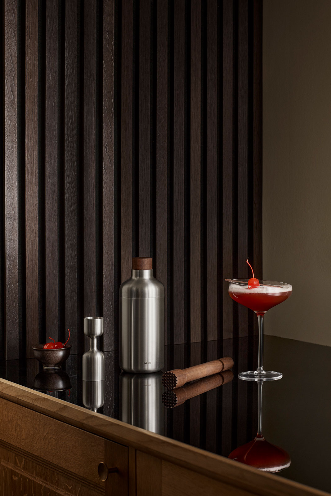 Ein Sideboard bei schwummerigen Licht. Auf der Ablage befinden sich der Cocktailshaker und weitere Artikel der Serie sowie ein gefülltes Cocktailglas.
