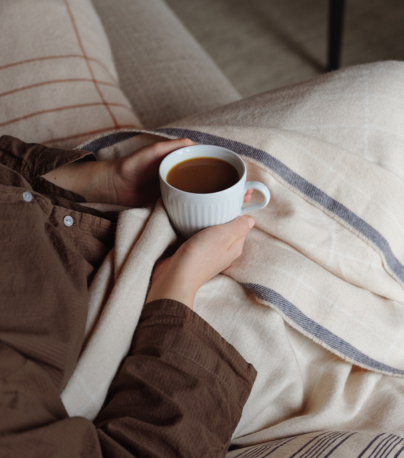 Zwei Frauenhände halten eine Legio Nova Kaffeetasse fest. Die Person sitzt in eine Decke gekuschelt auf dem Sofa.