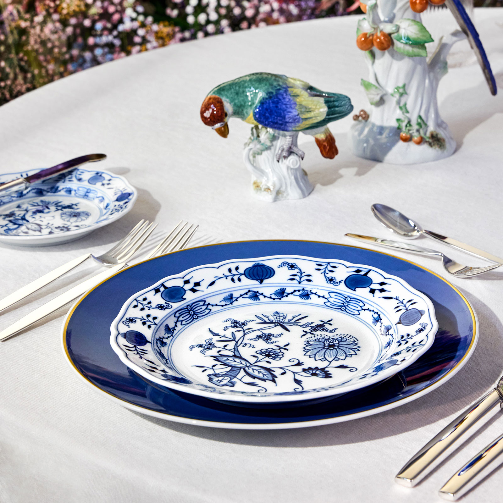 Auf einem gedeckten Tisch steht ein Teller im traditionellen Zwiebelmuster auf dem blauen Urban Colors Speiseteller von Meissen. Neugierig beugt sich ein kleiner bunter Vogel vor, um die Kombination zu betrachten.