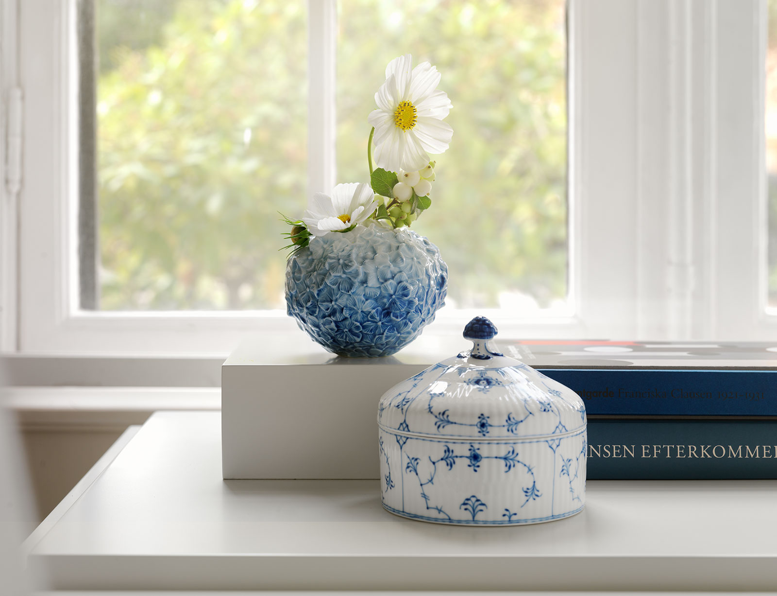 Mit einer weißen Blume und einem Zweig darin steht die Hortensien Vase hellblau auf einem dicken Buch vor einem hellen Fenster.